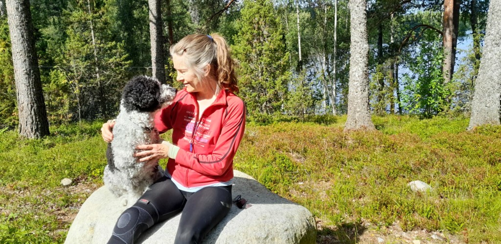 Elina Järvinen istuu kivellä punainen takki yllään. Elina katsoo vieressään olevaa koiraa. Taustalla on metsää. Aurinko paistaa