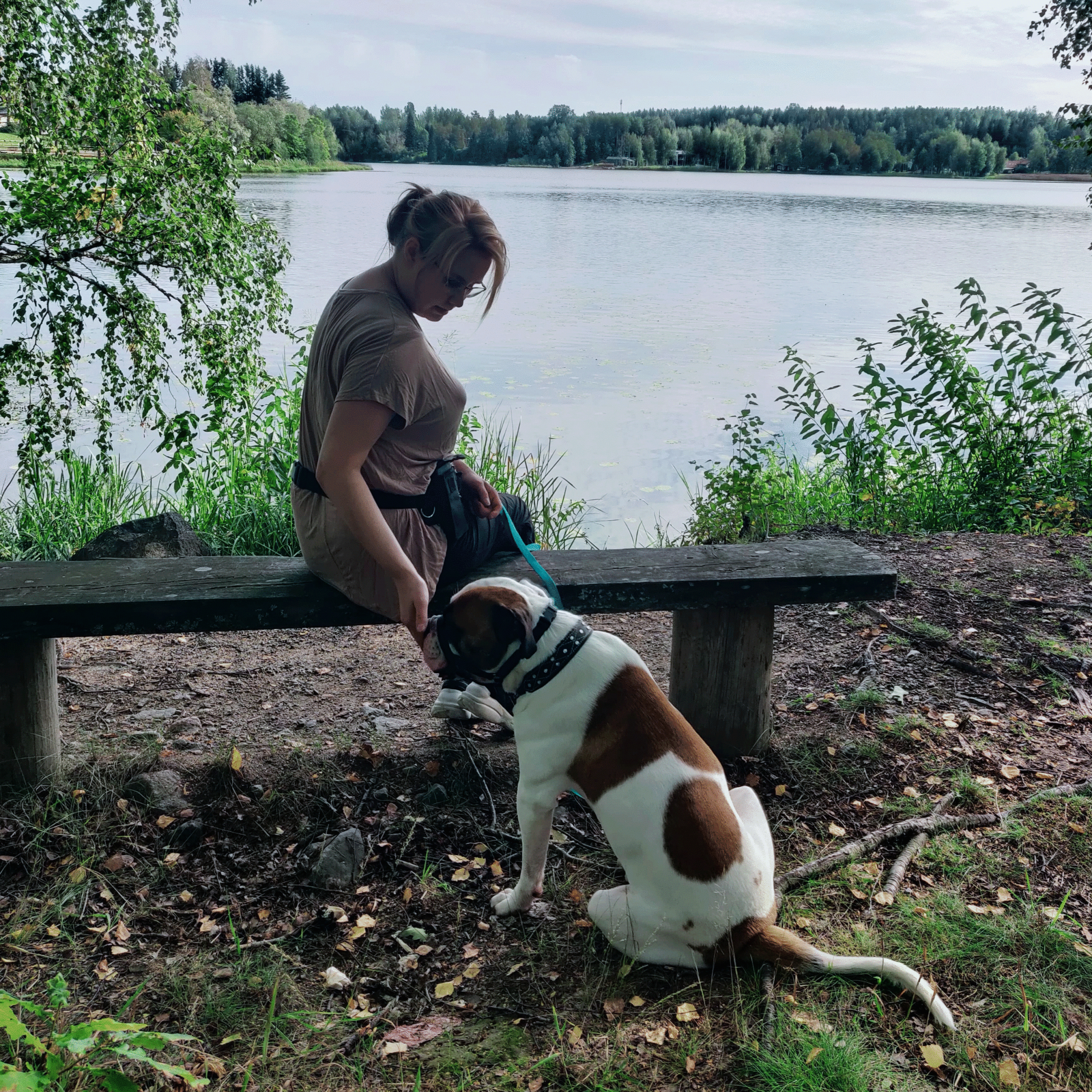 Maiju Moilanen istuu penkillä järven rannalla. Maijun vieressä maassa on ruskeavalkoinen koira, jota hän rapsuttaa.