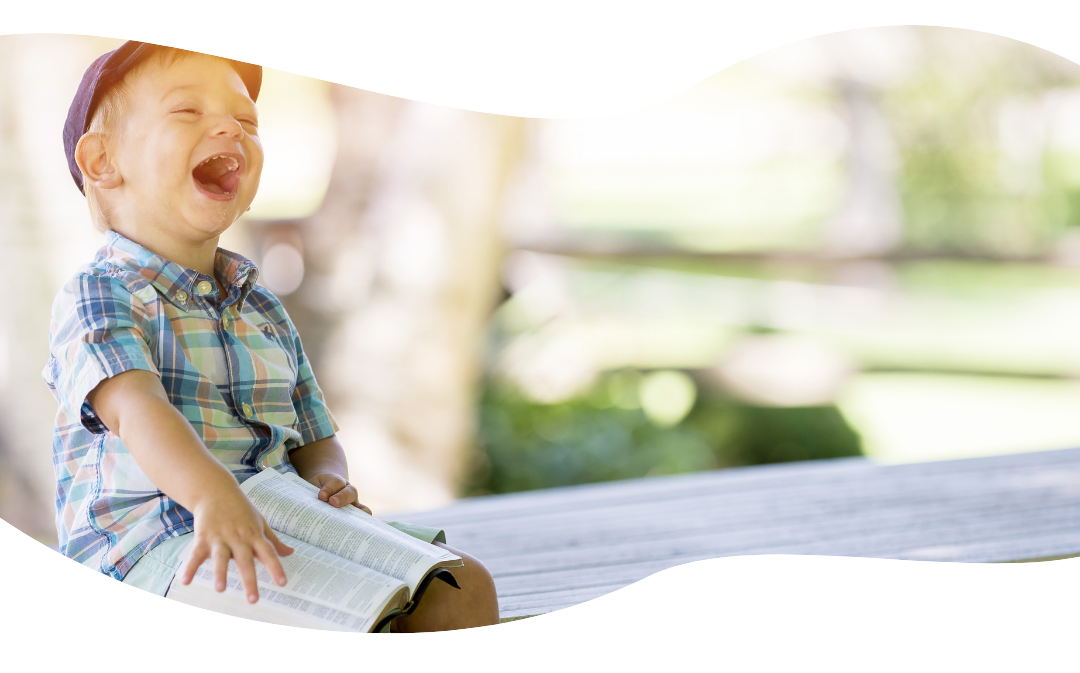 Kuvan vasemmassa reunassa istuu noin 3-vuotias poika nauraen. Pojalla on sylisssään kirja, jota hän selaa.