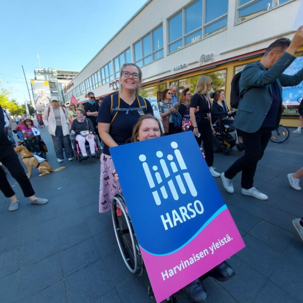 Kaksi naista osallistumassa Vammaisvoimaa mielenilmaukseen Helsingin keskustassa. Ympärillä on paljon muitakin ihmisiä. Toinen naisista istuu pyörätuolissa ja pitää käsissään kylttiä, jossa lukee isoin kirjaimin HARSO.