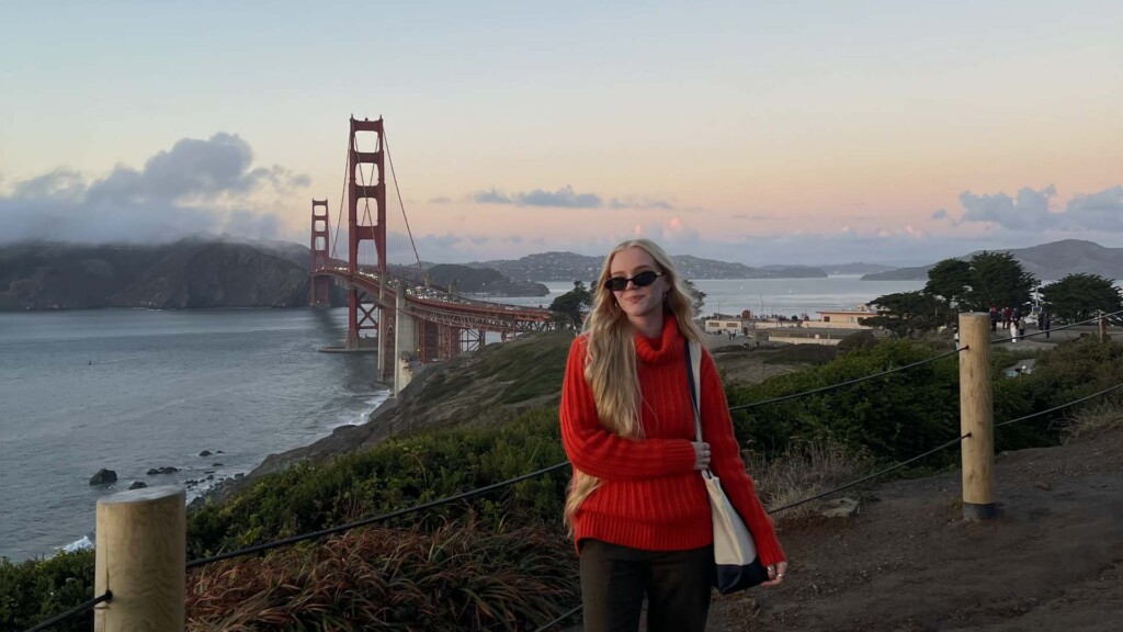 Iida on kuvan keskellä ja hänellä on päällään punainen paita ja mustat aurinkolasit. Kuvan taustalla näkyy San Fransiscon Golden Gate -silta auringinlaskussa.